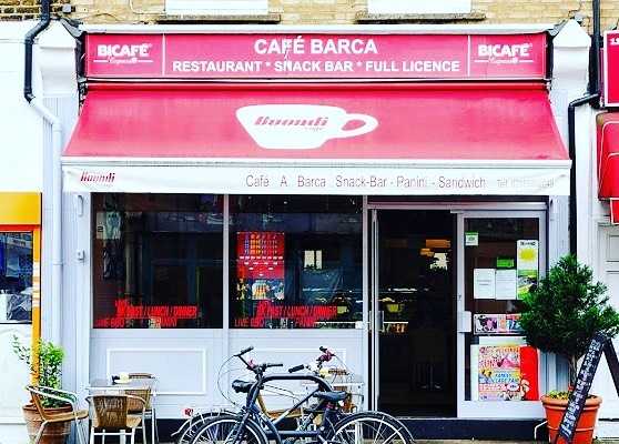 The front of Cafe Barca on Fulham Palace Road. A red awning over the window, and a sign above it reading CAFE BARCA, restaurant, snack bar, full licence. Two tables and chairs on the pavement, and bicycles parked at the kerb.